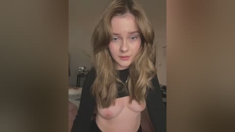 Video of a young Caucasian woman with long, wavy blonde hair, wearing a black top lifted to expose her bare breasts, standing in a dimly lit room with neutral-colored walls and a bed in the background.