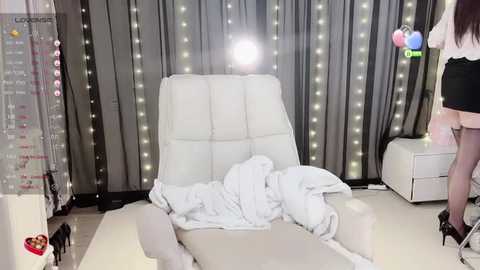 Video of a woman in a white dress with black high heels, standing beside a white chair draped with a white blanket, against a backdrop of gray curtains with fairy lights.