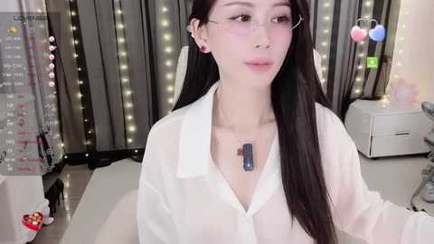 Media: Video of an East Asian woman with long black hair, fair skin, and a slender build, wearing a sheer white blouse, seated indoors with string lights and a calendar in the background.