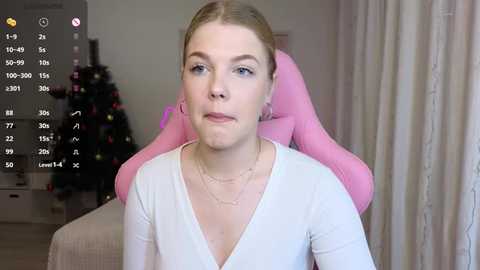 Video of a fair-skinned, blonde woman with blue eyes wearing a white V-neck top, seated in a pink gaming chair. The background features a Christmas tree and beige curtains in a well-lit room.