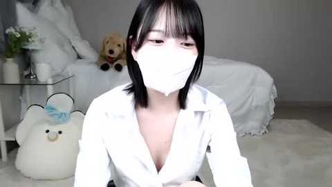 Media: Video of an Asian woman with straight black hair, wearing a white mask, white blouse, and a plush toy, seated on a fluffy white rug in a minimalistic, light-toned bedroom.