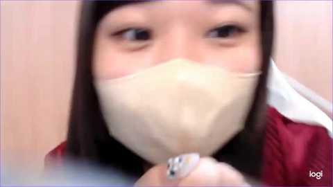 Media: A video of a young woman with straight, dark hair wearing a beige surgical mask, a white collar, and a red jacket, seen in close-up.