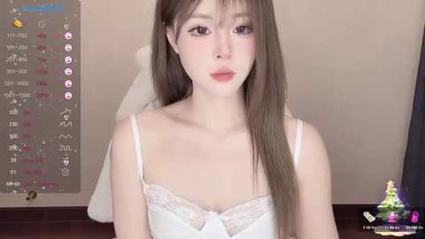 Media: Video of a young Asian woman with fair skin, long brown hair, and wearing a white lace-trimmed camisole. Background includes a beige wall with a calendar, and a screen with social media icons.