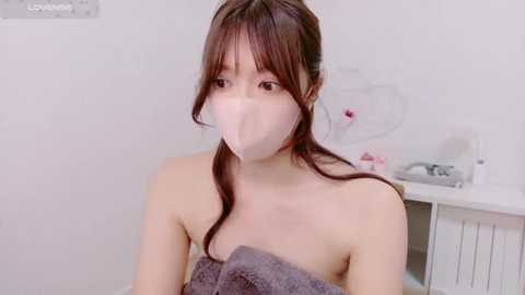 Media: A video of an East Asian woman with fair skin, long brown hair, and a white surgical mask covering her mouth. She is wrapped in a grey towel, standing in a white bathroom with a white radiator and a sink in the background.