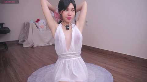 Media: Video of an Asian woman with long black hair, wearing a white, sleeveless, plunging neckline dress, sitting cross-legged on a round rug in a minimalist room with wooden floors, soft pastel decor, and a cat toy in the background.