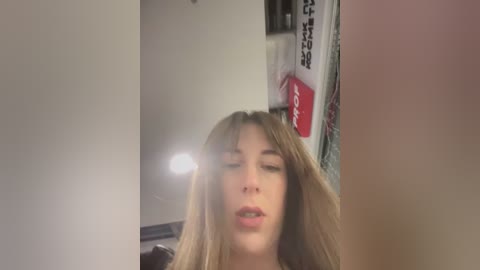 Media: A video of a young woman with long, light brown hair, fair skin, and a neutral expression, captured in a dimly lit room with a blurred background featuring a red sign and a white object.