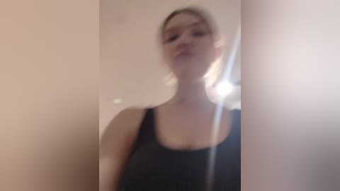 Media: A blurred video of a young Caucasian woman with light blonde hair, wearing a black tank top, standing in front of a mirror with a blurred background.