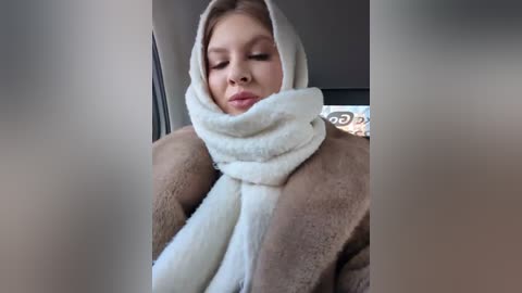 Media: Video of a fair-skinned woman with blonde hair, wearing a white scarf and a brown coat, sitting in a car, eyes closed, serene expression.