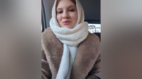 Media: Video of a fair-skinned woman with a light brown fur coat, wearing a white scarf and light makeup, sitting in a car. Background is blurred.