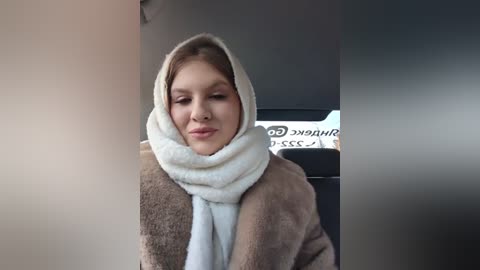 Media: Video of a smiling woman with fair skin and brown hair, wearing a white scarf and brown coat, inside a car with a \"Starbucks\" sign visible.