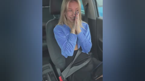 Media: Video of a blonde woman wearing a blue sweater and black skirt, seated in a car, holding a cell phone to her ear, with a seatbelt across her lap.