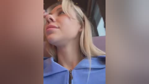 Media: Video of a young woman with straight blonde hair, wearing a blue hoodie, sitting in a car with blurred background, looking upward with a thoughtful expression.