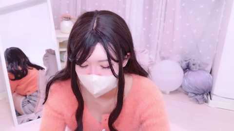 Media: Video of an Asian woman with long black hair, wearing an orange sweater and a white face mask, sitting in a room with white walls, polka-dot curtains, and fluffy pillows.