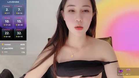 Media: Video of an East Asian woman with long black hair, wearing a black off-shoulder top, against a colorful, abstract background.