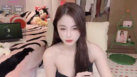 Media: Video of an East Asian woman with long, straight black hair, wearing a strapless black top, seated in a cozy room with a green stool, a plush Mickey Mouse, and a laptop.