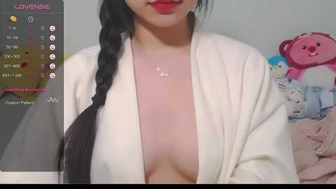 Media: A video of an East Asian woman with a light skin tone, wearing a white robe, exposing her left breast, with a braided black hair. Background shows plush toys.