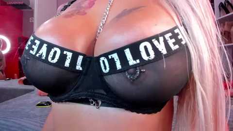 Media: Video of a woman with platinum blonde hair, wearing a black bra with \"I LOVE TO TAN\" written in white letters across the cups. Her large breasts are prominently displayed.