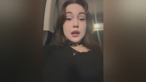 Media: Video of a young woman with fair skin and long, wavy brown hair, wearing a black ribbed top and a black choker necklace, sitting in a dimly lit car.