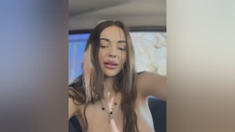 Media: Video of a young woman with long brown hair, fair skin, and small breasts, wearing a black top, taken from a low angle, showing a blurred background.