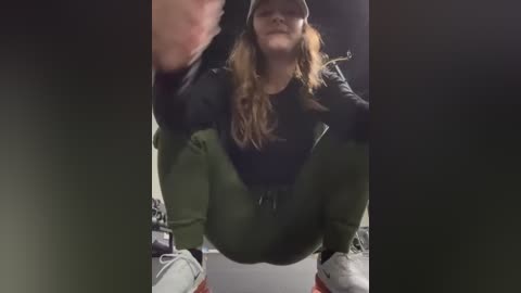 Media: Video of a young woman with long, blonde hair, wearing a black hoodie, green pants, and white sneakers, squatting in a dimly lit room.