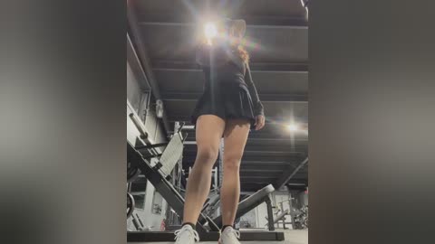 Media: Video of a woman in a black sports outfit, short skirt revealing legs, lifting weights in a gym, bright flash, industrial ceiling, blurred background.