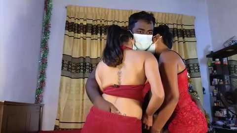 Media: Video of three women in a room with a yellow and brown curtain; one woman with a red dress, another in a red lace dress, and a third in a red dress, all wearing face masks.