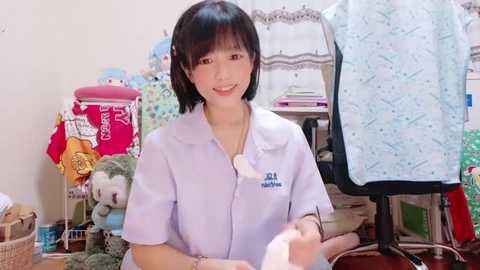 Media: A video of a young Asian woman with short black hair and a soft smile, wearing a white blouse, holding a baby doll. The background features a cluttered room with toys and clothes.