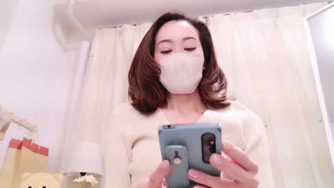 Media: Video of a light-skinned Asian woman with shoulder-length brown hair, wearing a beige knitted sweater, white face mask, and a blue smartphone. She's taking a selfie in a white-tiled bathroom with a cream-colored shower curtain.