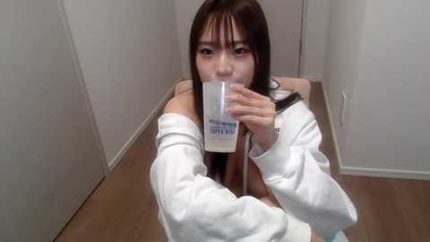 Media: A video of an Asian woman with long black hair, wearing a white jacket, drinking from a white cup in a minimalist, white-walled room with wooden floors.