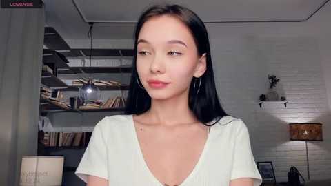 Video of an East Asian woman with long black hair, wearing a white V-neck shirt, standing in a modern, well-lit room with white brick walls, shelves, and a cozy lamp.