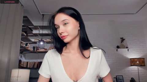 Video of a slender, light-skinned woman with straight black hair, wearing a white V-neck t-shirt, in a cozy, book-filled room with exposed brick walls and modern lighting.