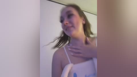 Media: A blurred video of a young woman with light skin, wearing a white spaghetti-strap top, standing indoors against a plain white wall, smiling slightly.