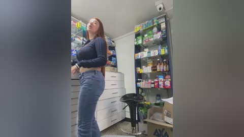 Media: Video of a tall, curvy Caucasian woman with long red hair, wearing a tight, dark blue crop top and high-waisted jeans, standing in a cluttered, brightly lit pharmacy.