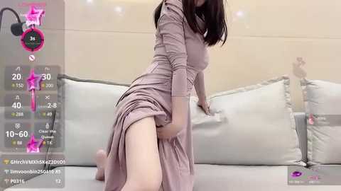 Media: Video of a fair-skinned woman with dark hair in a short dress, kneeling on a bed with white pillows, capturing a playful and suggestive moment.