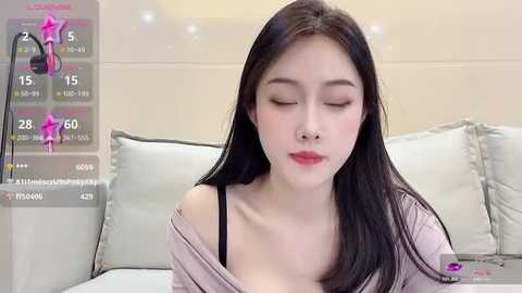 Media: Video of an Asian woman with long black hair, wearing a light pink off-shoulder top, sitting on a white couch, eyes closed, looking serene.