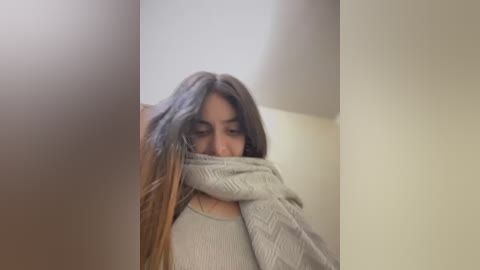 Media: A video of a young woman with long, straight brown hair, wearing a light gray, ribbed sweater, partially covering her face with the sweater's arms. The background is a plain, beige wall.