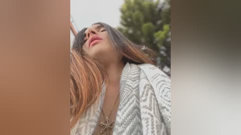 Media: Video of a woman with long, straight, brown hair, wearing a white patterned robe, standing outdoors with a blurred green tree in the background.