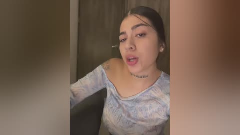 Media: A video of a Latina woman with medium skin tone, dark hair, and a choker necklace, wearing a light-colored off-shoulder top. She has a shocked expression, with a blurred background featuring a wooden door.