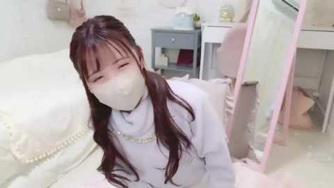 Media: A video of a young girl with long dark hair, wearing a white mask and white clothes, kneeling on a bed in a messy room with a pink mirror.