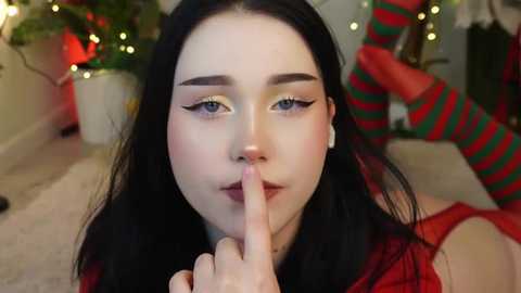 Media: A video of a fair-skinned woman with long black hair, wearing a red dress, and black eyeliner, covering her mouth with her finger, against a festive background with green and red striped stockings and potted plants.