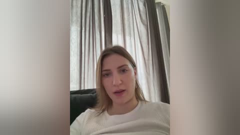 Media: Video of a young woman with straight, shoulder-length brown hair, wearing a white shirt, sitting on a black couch in a dimly lit room with sheer white curtains and dark brown drapes.