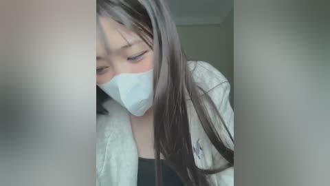 Media: Video of an Asian woman with long dark hair, wearing a white face mask and a light jacket, bending over to look at a blurred object.