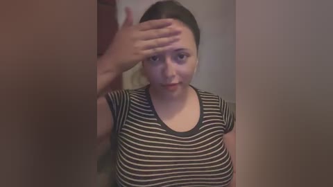 Media: Video of a young woman with fair skin and dark hair, wearing a black-and-white striped shirt, covering her forehead with her hand, indoors.