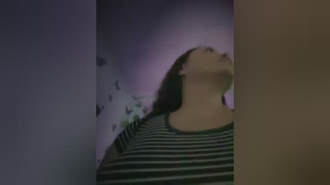 Media: Video of a young woman with long brown hair, wearing a green and white striped shirt, standing in a dimly lit room with a blurred, mysterious background.