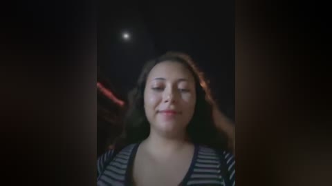 Media: Video of a young woman with long, wavy brown hair, wearing a striped shirt, standing in a dimly lit room. Her eyes are closed, and she appears to be smiling.