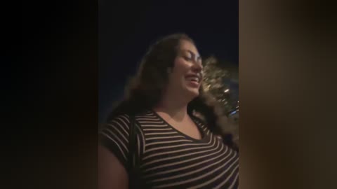 Media: Video of a smiling, curvy, light-skinned woman with long brown hair, wearing a black and white striped top, standing in a dimly lit room with blurred background.