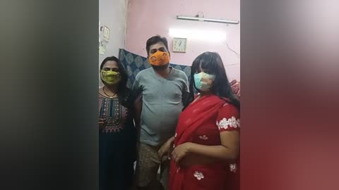 Media: Video of three people in a dimly lit room, wearing masks. A man in a gray shirt stands in the middle, flanked by a woman in a red sari and another woman in a blue dress.