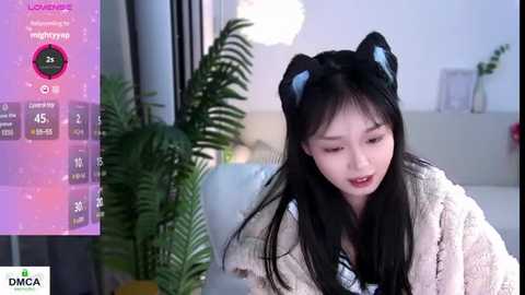 Media: Video of an Asian woman with long black hair wearing cat ears and a white knitted sweater, smiling in a modern, brightly lit room with a green plant and digital clock in the background.