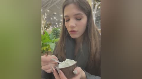 Media: Video of a young woman with long brown hair, wearing a black top, eating ice cream from a waffle cone, in a greenhouse setting with lush greenery and string lights.