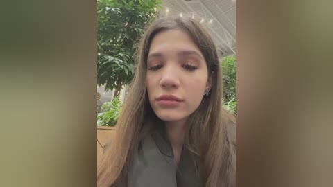 Media: A video of a young woman with long brown hair and light skin, wearing a green shirt, in a lush indoor garden with green plants.